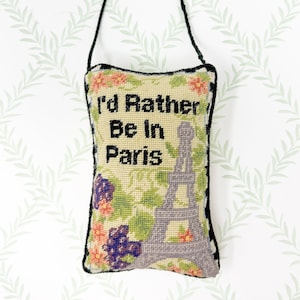 May include: A rectangular, hand-stitched pillow with the text "I'd Rather Be In Paris" and an Eiffel Tower illustration. The pillow features floral and vine patterns in shades of pink, purple, and green, with a black border and a black handle.