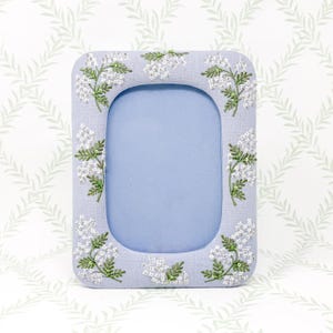 May include: A rectangular photo frame with a pale blue fabric border. The frame is decorated with embroidered white flowers and green leaves. The inner frame is a light blue colour.