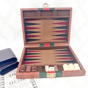 Könnte beinhalten: Offenes Backgammon-Set mit braunem Lederetui. Das Spielbrett hat abwechselnd rote, schwarze und weiße Dreiecke. Enthält braune und weiße Spielsteine, Würfel und einen Würfelbecher. Das Etui hat grüne und rote Akzente.