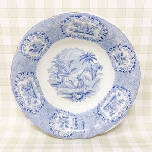 May include: A round ceramic bowl featuring a blue and white transferware pattern. The center depicts a landscape with palm trees and a figure, while the rim is decorated with alternating floral and architectural vignettes on a textured background.