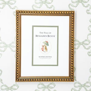 May include: Framed print of "The Tale of Benjamin Bunny" by Beatrix Potter. The print features a gold frame with a beaded edge, surrounding a white mat and a green border around the book cover illustration. The background is a white wall with green bow patterns.