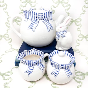 Peut inclure: Une théière, un pot à crème et un sucrier blancs avec des nœuds rayés bleus et blancs. Un lapin en céramique blanc avec un nœud rayé bleu et blanc est également inclus. L'ensemble est posé sur une pile de livres.