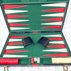 May include: A backgammon board game set featuring a green felt interior with alternating red and cream-coloured triangular points. The set includes two black leather dice cups, a stack of red checkers, a stack of cream checkers, and three white dice.