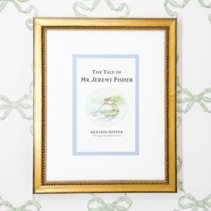 Może przedstawiać: Oprawiony wydruk "The Tale of Mr. Jeremy Fisher" autorstwa Beatrix Potter. Grafika przedstawia żabę na liściu lilii wodnej. Wydruk umieszczono w jasnoniebieskim passe-partout i złotej ramie z koralikową krawędzią. Tło to biała ściana z zielonym wzorem kokard.