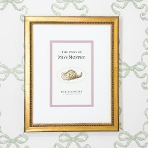 May include: A framed print of "The Story of Miss Moppet" by Beatrix Potter. The print features a light brown cat illustration on a pink background, set within a gold-colored frame. The background is a white wall with a repeating pattern of green bows.