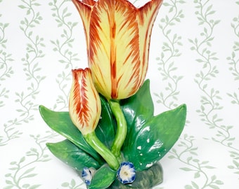 May include: A ceramic vase shaped like two tulips with yellow and red petals, supported by green leaves and stems. The base features small blue flowers on a dark green mound. The vase has a glossy finish and a sculptural, three-dimensional form.