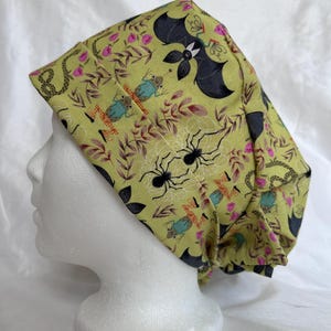 Puede incluir: Un gorro quirúrgico verde claro con un estampado temático de Halloween. El diseño de la tela incluye murciélagos negros, arañas, serpientes y ranas, junto con flores rosas y hojas marrones. El gorro está diseñado para ser usado en la cabeza.