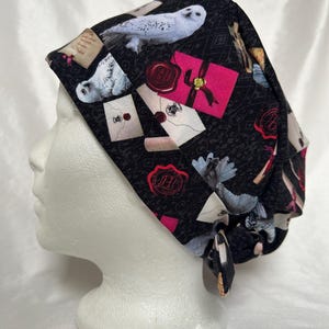 Può includere: Cappello chirurgico nero con un motivo ripetuto di gufi bianchi, buste rosa e pergamene. Il cappello ha una chiusura con lacci. Il design include sigilli di cera rossi e un motivo ripetuto.