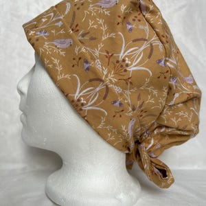 May include: Mustard-coloured scrub cap featuring a pattern of birds, leaves, and flowers in white and purple. The cap is tied at the back and is displayed on a white mannequin head.