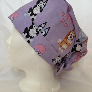 Puede incluir: Un gorro de quirófano de color morado claro con un patrón repetitivo de perros de dibujos animados en negro, blanco y naranja, junto con formas florales rosas. El gorro tiene una banda frontal doblada y un cierre de lazo en la parte posterior.