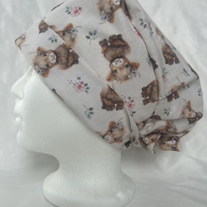 May include: A light beige scrub cap with a repeating pattern of brown Highland cows and floral accents. The cap is designed to cover the head and hair, often used in medical or culinary settings.