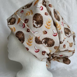 Könnte beinhalten: Eine OP-Haube mit hellbeigem Hintergrund und einem wiederkehrenden Muster aus braunen Igeln, roten Pilzen, Eicheln und gelben Herbstblättern. Die Kopfbedeckung aus Stoff hat ein gefaltetes Stirnband und Bänder zum Binden am Hinterkopf.