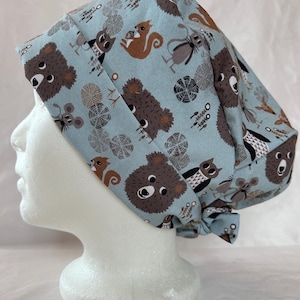 May include: A light blue scrub hat with a woodland animal print featuring bears, squirrels, owls, and mice. The hat has a tie at the back.