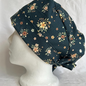 Puede incluir: Un gorro quirúrgico de color verde oscuro con un patrón floral repetitivo de pequeñas flores amarillas, naranjas y blancas con hojas verdes. El gorro tiene una banda ancha doblada en la parte delantera y se ata en la parte posterior con cintas.