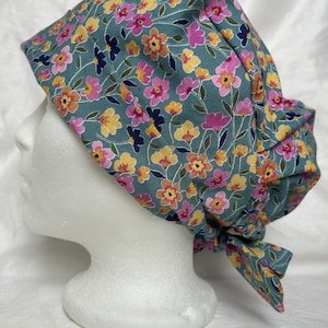 May include: A teal floral scrub cap with a pattern of pink, yellow, and orange flowers and navy blue accents. The cap is tied at the back and is displayed on a white mannequin head.