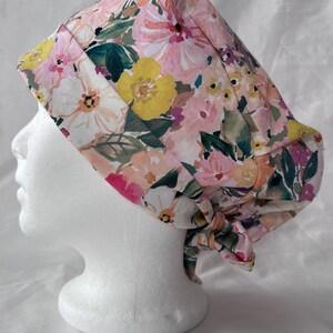 May include: A floral scrub cap with a watercolour design of pink, yellow, and white flowers and green leaves. The cap is tied at the back and displayed on a white mannequin head.