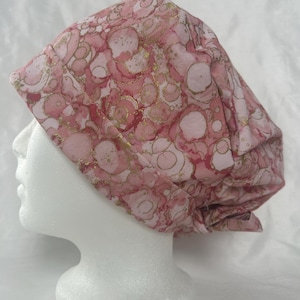 May include: A pink and gold patterned scrub cap. The cap is made of fabric with a design of pink circles and gold accents. The cap is displayed on a white mannequin head.