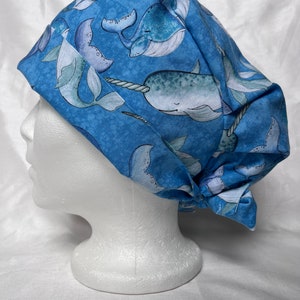 May include: A blue scrub hat with a pattern of narwhals and their tails. The hat is tied in a knot at the back.