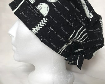 Op de afbeelding: Zwarte scrub-muts met een witte anatomische illustratie van een schedel, botten en skeletkenmerken. De tekst bevat "Sphenoid bone", "Floating Ribs", "Mental tuberosity", "Thoracic vertebrae", "Radius" en "Calcaneus".