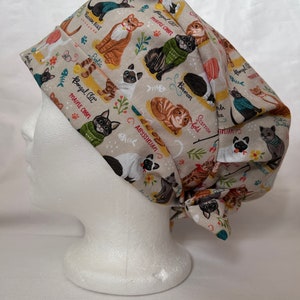 Könnte beinhalten: Eine beige OP-Mütze mit einem bunten Muster aus Katzen und ihren Rassenamen. Die Mütze hat einen Knoten am Hinterkopf.