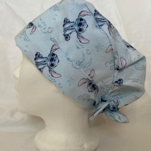 Puede incluir: Un gorro quirúrgico de color azul claro con un patrón repetitivo del personaje Stitch de Lilo y Stitch, intercalado con motivos de pequeñas estrellas de mar y olas. El gorro tiene una banda frontal plegada y un cierre de lazo en la nuca.