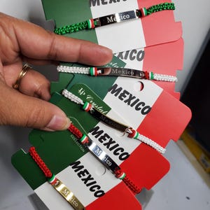 Puede incluir: Varios brazaletes con la palabra "México" en una placa de metal. Los brazaletes tienen detalles rojos, blancos y verdes, y se exhiben en tarjetas con la bandera mexicana. Los brazaletes están hechos de cordón tejido.