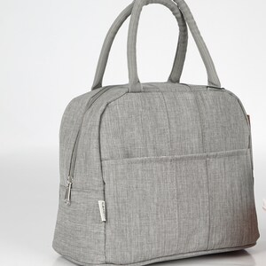 Puede incluir: Una bolsa de almuerzo aislada gris con cierre de cremallera y asa superior. La bolsa tiene un peque&ntilde;o bolsillo en la parte delantera con la palabra "CAMINO" impresa en ella.