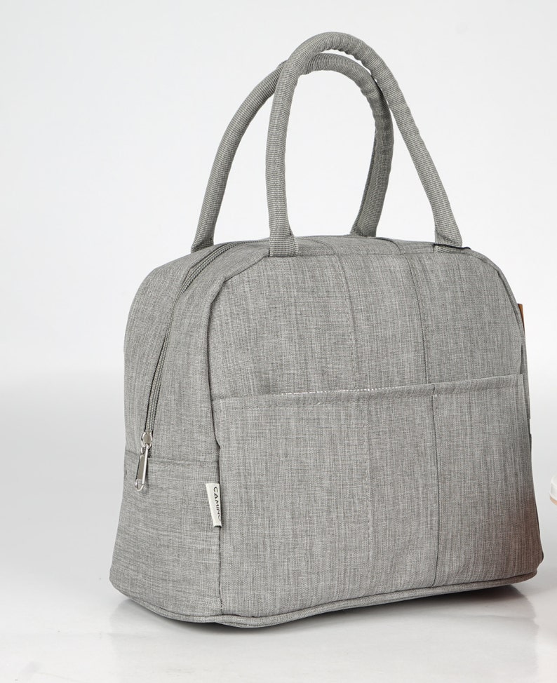 Puede incluir: Una bolsa de almuerzo aislada gris con cierre de cremallera y asa superior. La bolsa tiene un peque&ntilde;o bolsillo en la parte delantera con la palabra "CAMINO" impresa en ella.