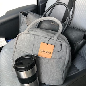 Puede incluir: Una mochila gris con una etiqueta marr&oacute;n que dice "CAMINO todo el tiempo contigo" y una taza de viaje negra y plateada. La mochila est&aacute; sentada en el asiento trasero de un coche con dos bolsos negros.