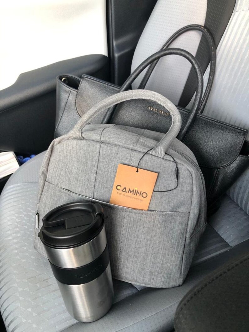 Puede incluir: Una mochila gris con una etiqueta marr&oacute;n que dice "CAMINO todo el tiempo contigo" y una taza de viaje negra y plateada. La mochila est&aacute; sentada en el asiento trasero de un coche con dos bolsos negros.