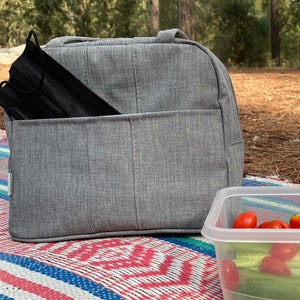 Puede incluir: Una bolsa de almuerzo gris con una mascarilla negra en el bolsillo delantero. La bolsa est&aacute; sentada sobre una manta de picnic colorida junto a un recipiente de pl&aacute;stico transparente con tomates rojos y pepinos verdes.