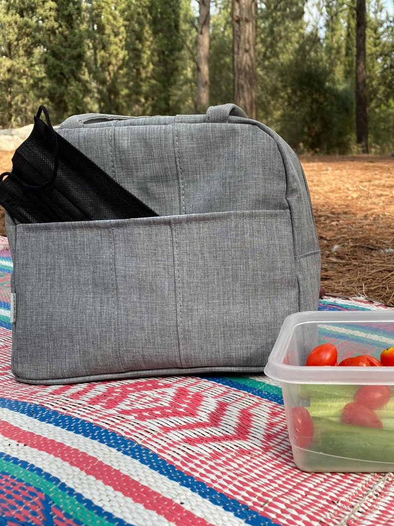Puede incluir: Una bolsa de almuerzo gris con una mascarilla negra en el bolsillo delantero. La bolsa est&aacute; sentada sobre una manta de picnic colorida junto a un recipiente de pl&aacute;stico transparente con tomates rojos y pepinos verdes.