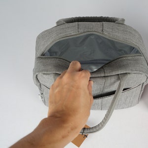 Puede incluir: Una bolsa de almuerzo aislada gris con una parte superior con cremallera y un forro de malla. La bolsa tiene un solo asa y un peque&ntilde;o bolsillo en el lateral.