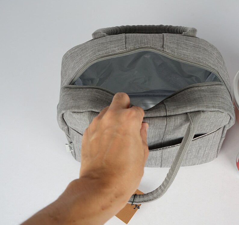 Puede incluir: Una bolsa de almuerzo aislada gris con una parte superior con cremallera y un forro de malla. La bolsa tiene un solo asa y un peque&ntilde;o bolsillo en el lateral.