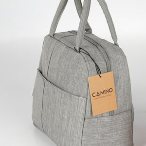 Puede incluir: Una bolsa de almuerzo gris con cierre de cremallera y un peque&ntilde;o bolsillo lateral. La bolsa tiene una etiqueta marr&oacute;n con el texto "CAMINO todo el tiempo contigo".