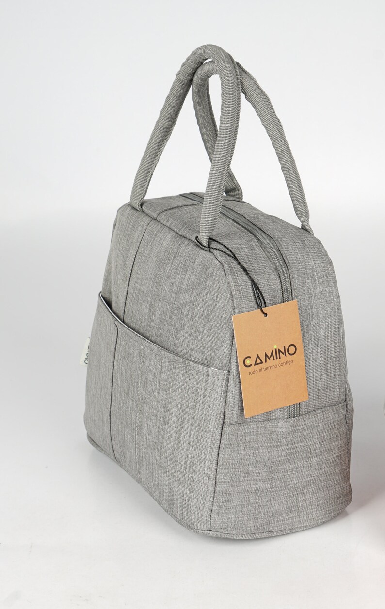 Puede incluir: Una bolsa de almuerzo gris con cierre de cremallera y un peque&ntilde;o bolsillo lateral. La bolsa tiene una etiqueta marr&oacute;n con el texto "CAMINO todo el tiempo contigo".