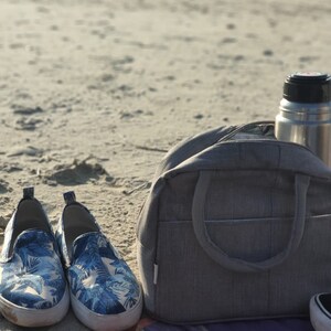 Puede incluir: Una bolsa de lona gris con una botella de agua plateada en la parte superior. La bolsa est&aacute; sentada en una playa de arena con un par de zapatos de tela azul y blanco con estampado floral delante.