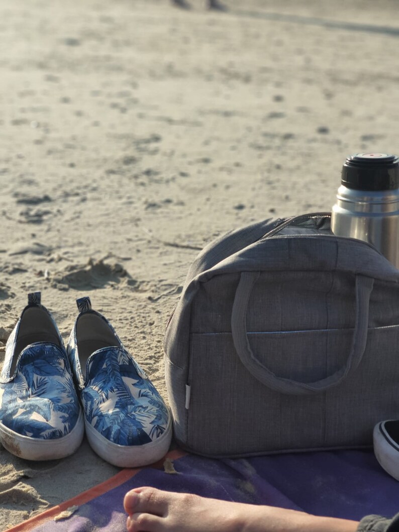 Puede incluir: Una bolsa de lona gris con una botella de agua plateada en la parte superior. La bolsa est&aacute; sentada en una playa de arena con un par de zapatos de tela azul y blanco con estampado floral delante.