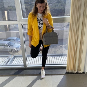 Puede incluir: Una mujer con un blazer amarillo, una camiseta blanca con el texto "Babe Don't Cry", pantalones vaqueros negros y zapatillas blancas. Lleva un bolso de mano gris.