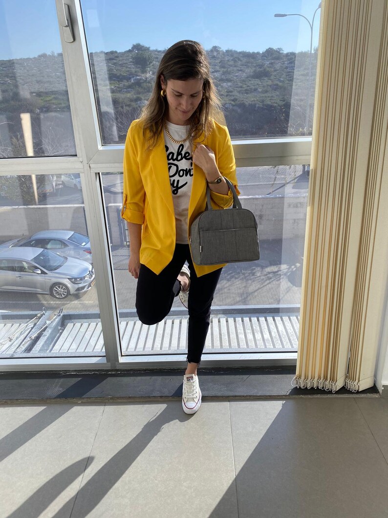 Puede incluir: Una mujer con un blazer amarillo, una camiseta blanca con el texto "Babe Don't Cry", pantalones vaqueros negros y zapatillas blancas. Lleva un bolso de mano gris.