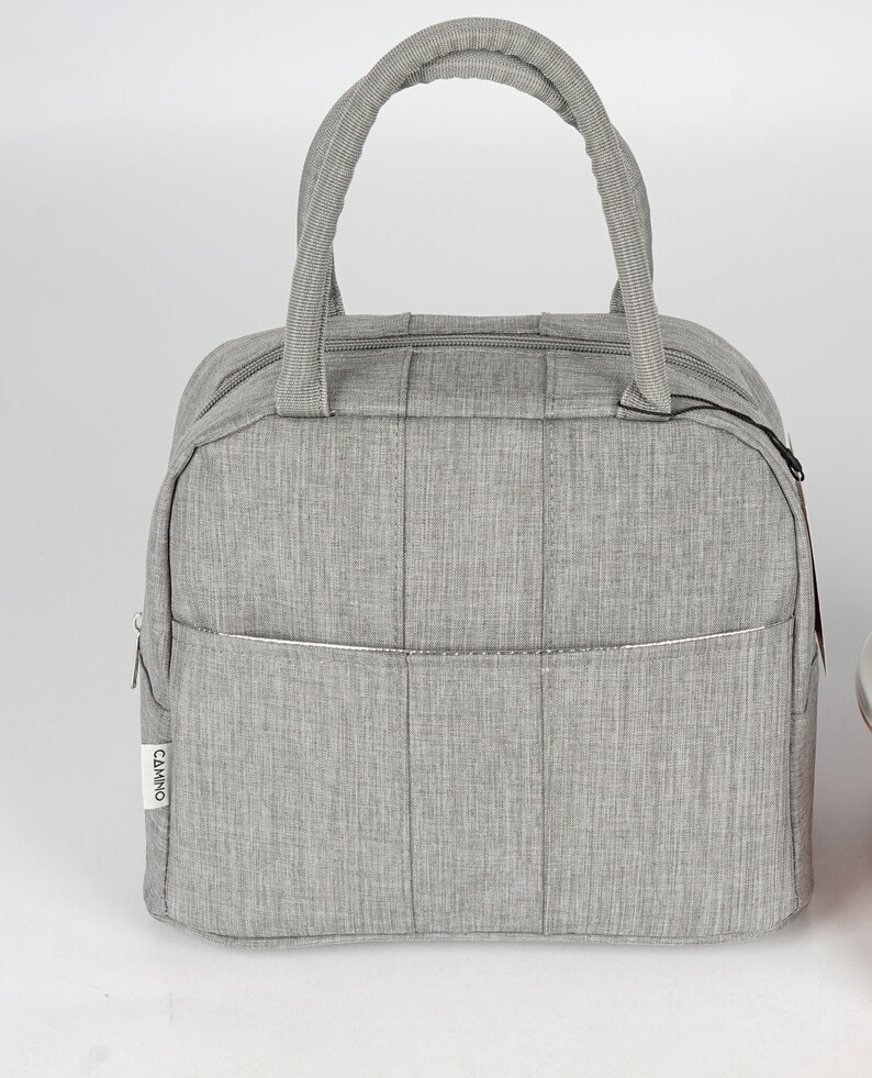 Puede incluir: Una bolsa de almuerzo gris con cierre de cremallera y asa superior. La bolsa tiene un peque&ntilde;o bolsillo en la parte delantera con la palabra "CAMINO" impresa en &eacute;l.