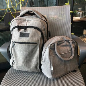 Puede incluir: Una mochila gris con una cremallera negra y la palabra "Matein" en la parte delantera. Junto a ella hay una bolsa de almuerzo gris m&aacute;s peque&ntilde;a con un asa negra.