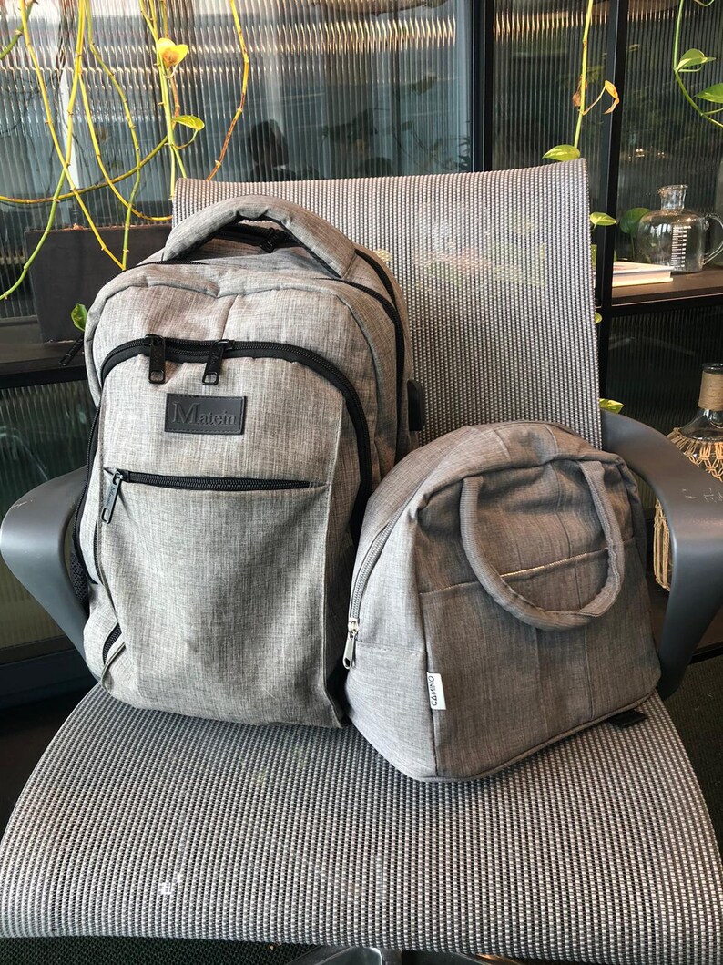 Puede incluir: Una mochila gris con una cremallera negra y la palabra "Matein" en la parte delantera. Junto a ella hay una bolsa de almuerzo gris m&aacute;s peque&ntilde;a con un asa negra.