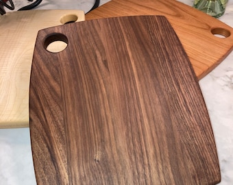 Peut inclure: Plusieurs planches à découper en bois de différentes nuances de brun. Les planches ont des coins arrondis et un trou circulaire pour les suspendre. Une bouteille de vin rouge et une bouteille de vin blanc sont en arrière-plan.