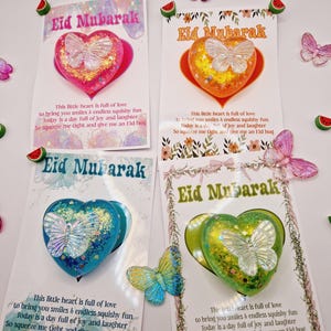 Pode incluir: Quatro ornamentos em forma de coração coloridos com glitter e uma borboleta dentro. Cada ornamento tem um cartão anexado com o texto "Eid Mubarak" e uma mensagem sobre amor, alegria e risos.