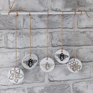 May include: Five white ceramic ornaments with black and gold bee designs hang from a white wooden dowel rod. The ornaments are suspended by twine.