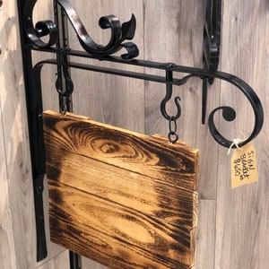May include: A rustic, wooden sign with a burnt wood finish hangs from a decorative black metal bracket. The bracket features scrolled accents and a hanging chain. The sign is rectangular and appears to be handmade.