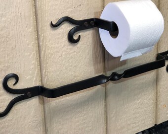 May include: Black wrought iron toilet paper holder and towel bar set. The toilet paper holder has a decorative scroll design, and the towel bar features a twisted center. The set is mounted on a light-colored wooden surface.
