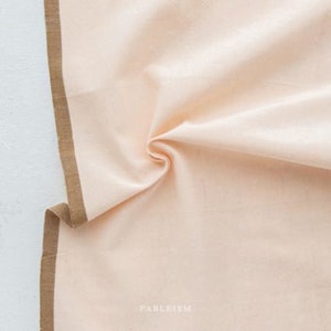 May include: A close-up of a light peach-colored fabric with a subtle texture. The fabric has a brown border along the edge. The text "FABLEISM" is printed in the bottom right corner.