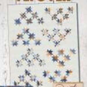 May include: A quilt with a repeating star pattern in shades of blue, tan, and white. The quilt features the text "FLY OVER" at the top and the designer's name at the bottom. The quilt is displayed on a white background.